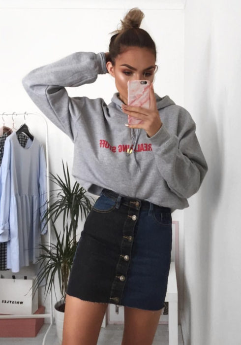 Looks con sudadera; mujer tomándose selfie frente al espejo, peinado de chongo alto, hoodie gris, falda de mezclilla