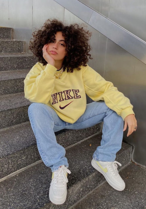 Looks con sudadera; mujer morena sentada en las escaleras, cabello chino esponjado, hoodie amarillo pastel, pantalón mom jean azul, tenis blancos