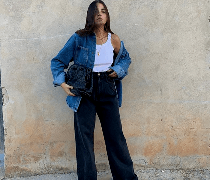 chica de cabello castaño usando un tank top blanco, camisa de mezclilla, pantalones acampanados de mezclilla y bolso azul marino de piel