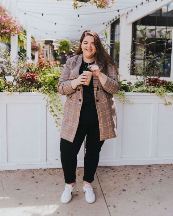 Chica curvy usando zapatos grises, blusa y pantalón negros y blazer color arena largo