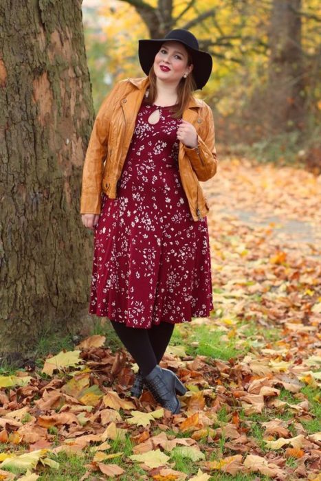 Chica curvy usando botines grises, medias y sombrero negros, vestido midi color vino con estampado floral y chaqueta mostaza