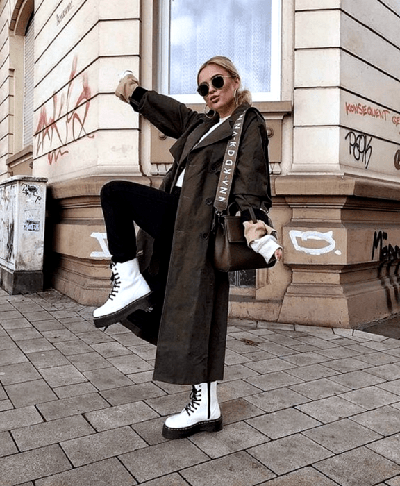 Chica rubia usando lentes de sol, camiseta blanca, leggings negros, botines blancos de plataforma negra, abrigo largo café oscuro y bolso café de piel
