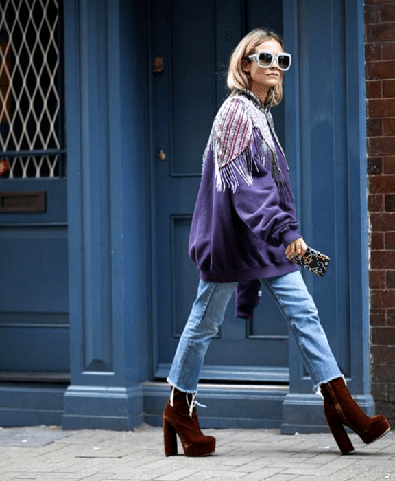 Chica rubia usando lentes de sol, sudadera morada con aplicaciones en los hombros, jeans cropped y zapatos de plataforma rojos de terciopelo