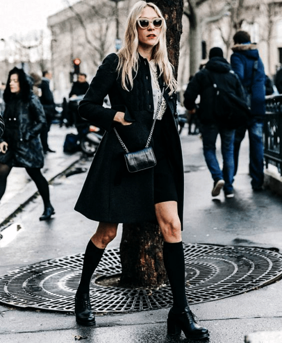 Chica rubia usando lentes de sol, camisa blanca, abrigo negro, minifalda negra, calcetas negras y zapatos de plataforma