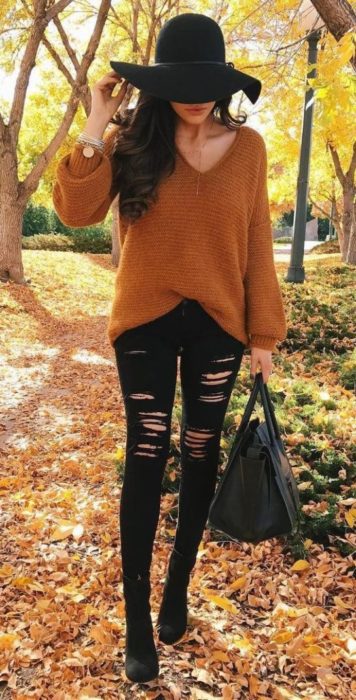 Chica usando jeans negros rasgados, botines negros, suéter color mostaza holgado, y sombrero