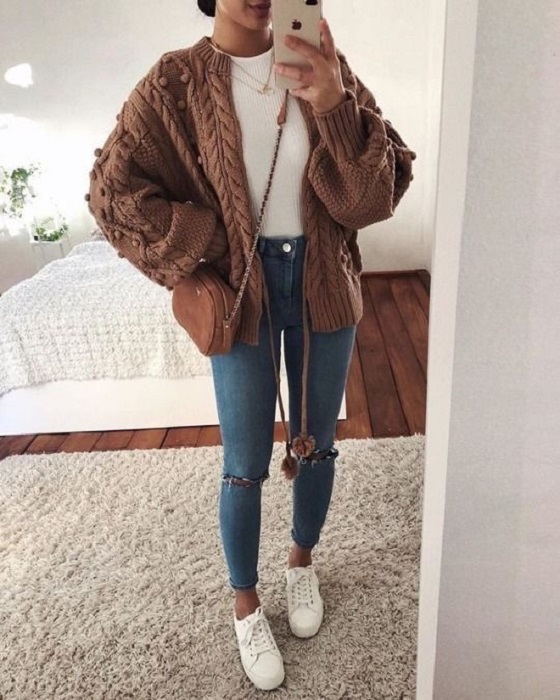Chica usando jeans rasgados, tenis blancos, top blanco y suéter tejido color café, con una bolsa de mano camel