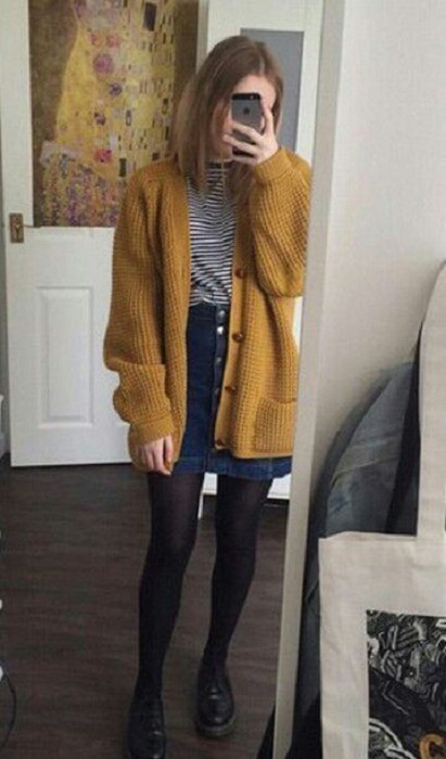 Chica usando botines, medias negras, falda denim, top gris y suéter largo color mostaza