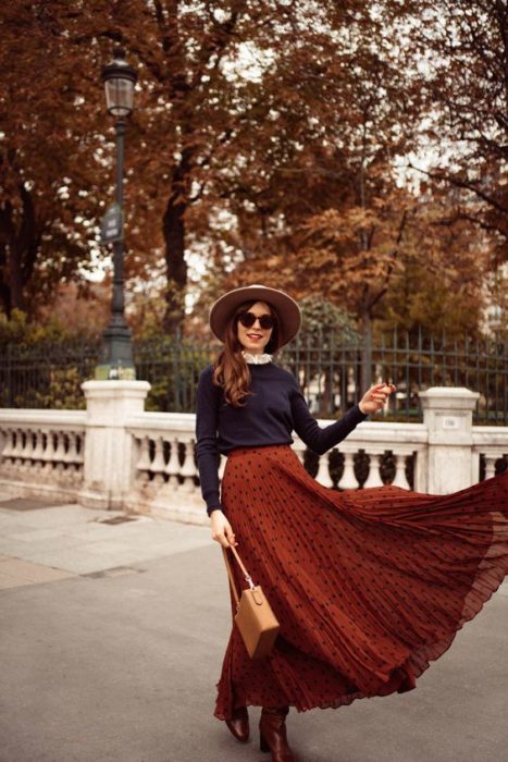 Chica de cabello largo y suelto castaño con gafas de sol y sombrero, usando suéter azul marino y maxi falda naranja