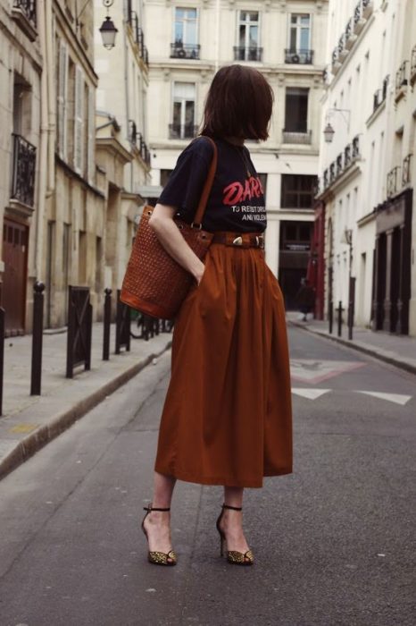 Chica posando en la calle con blusa negra y falda larga color naranja y tacones marrones