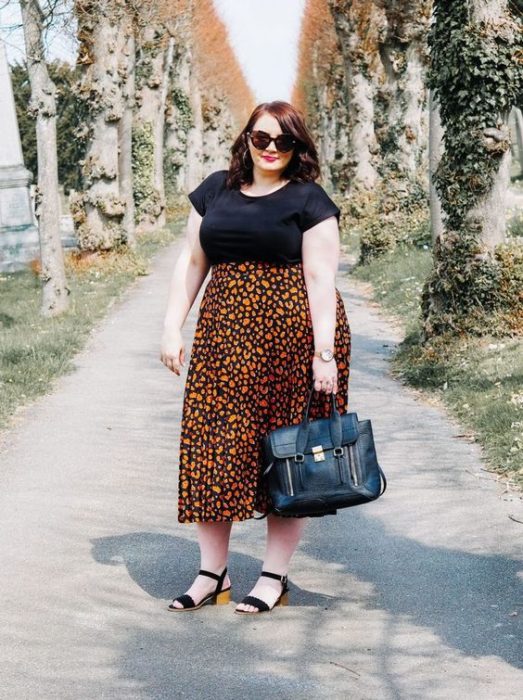 Chica plus size pelirroja con blusa negra y maxi falda naranja de flores y sandalias negras