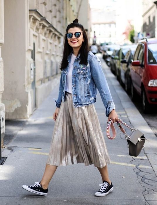 Chica caminando por la calle con falda metálica gris, Converse negros, blusa blanca y chaqueta de mezclilla