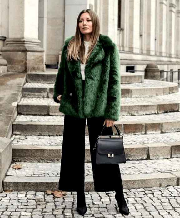 chica rubia usando camiseta blanca, abrigo afelpado verde, pantalones de vestir negros, botines de tacón negros y bolso negro
