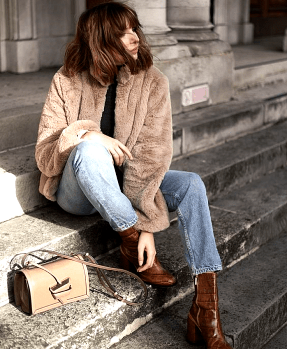 chica castaña usando abrigo afelpado marrón, top negro, jeans azules, botines de piel marrón de tacón, bolso de mano beige