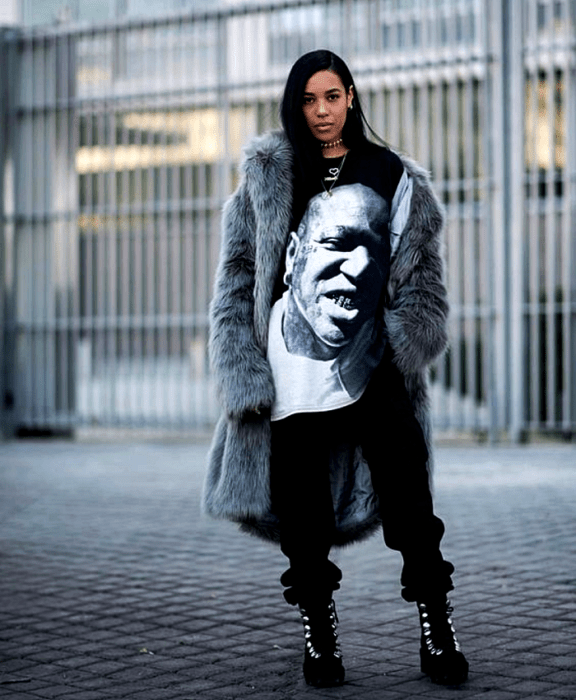 chica morena con cabello oscuro usando camiseta oversized negra con estampado, abrigo afelpado gris, pantalones negros holgados, y tenis de plataforma negros