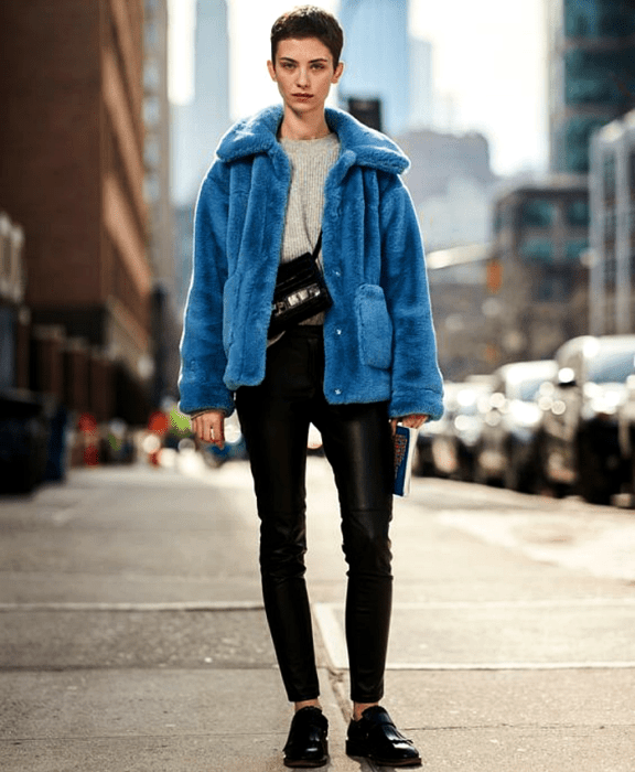 chica de cabello corto usando suéter gris, abrigo afelpado azul, bolso negro crossover, pantalones de cuero negros, y zapatos loafers negros