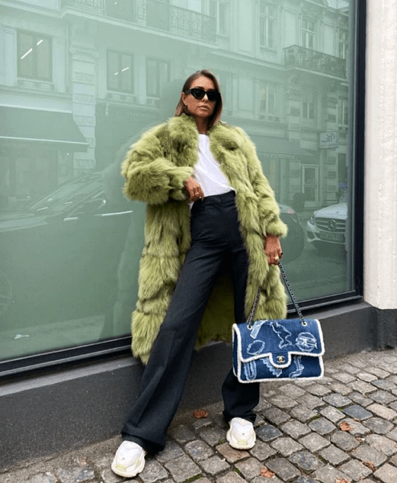 chica rubia usando lentes de sol, camiseta blanca, pantalones de vestir gris oscuro, tenis deportivos blancos, bolso de mezclilla grande, y abrigo afelpado verde