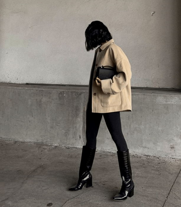 chica de cabello oscuro corto usando una chaqueta oversized beige, bolso mini de mano negro, leggings negros y botas largas de tacón de piel