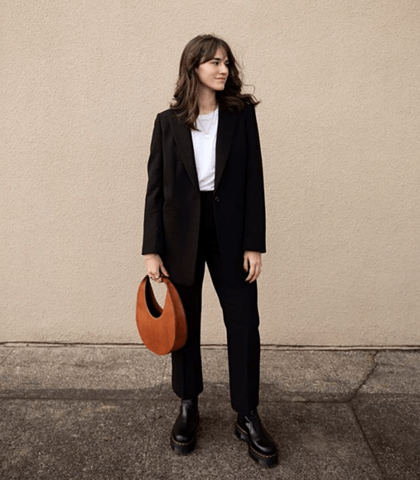chica de cabello castaño usando una camiseta blanca, blazern negro, pantalones de vestir negros, botas de suela gruesa negras y bolso de mano naranja