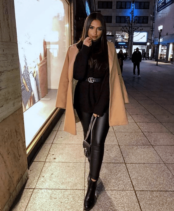 mujer de cabello largo con suéter negro, cinturón negro de piel, abrigo beige, leggings de cuero negro, botines negros de tacón y bolso negro
