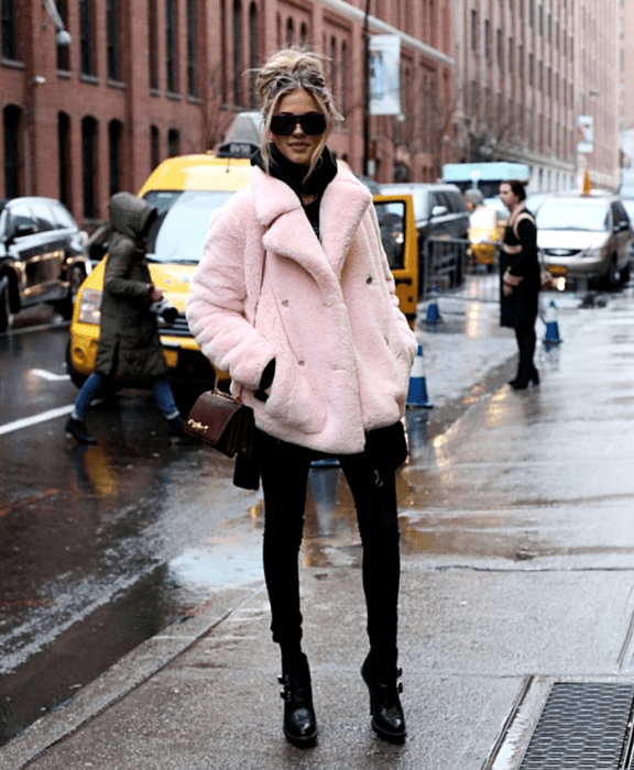 mujer rubia usando lentes de sol, abrigo furry rosa, top negro, leggings negros, botines de tacón negros y bolso mini de cuero guinda