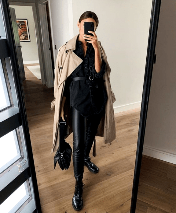 mujer de cabello castaño usando un abrigo largo marrón, camisa de botones negra, cinturón negro, leggings de cuero negro, botines de piso y bolso negro
