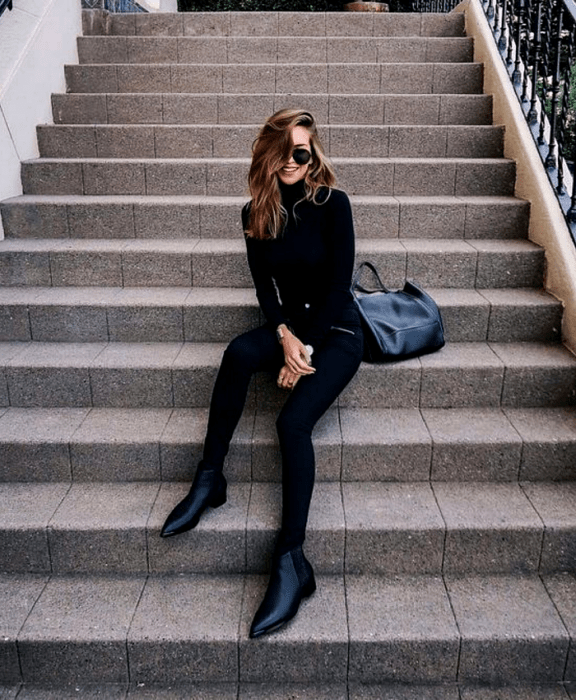 mujer de cabello claro usando lentes de sol, top negro de manga larga y cuello alto, leggings negros y botines de cuero sin tacón, bolso negro