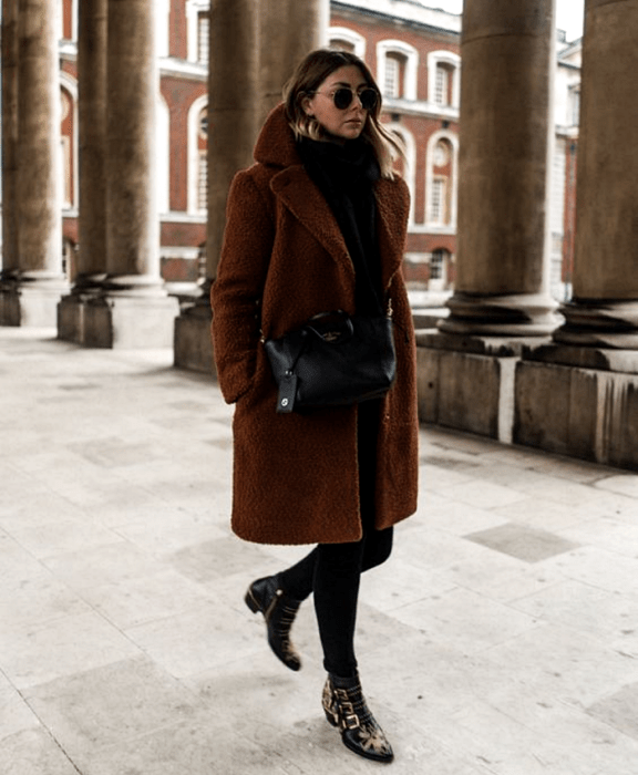 mujer de cabello claro con lentes de sol, abrigo teddy marrón, suéter negro de cuello alto, leggings negros, botines de piso con estampado de animal print y bolso negro de cuero