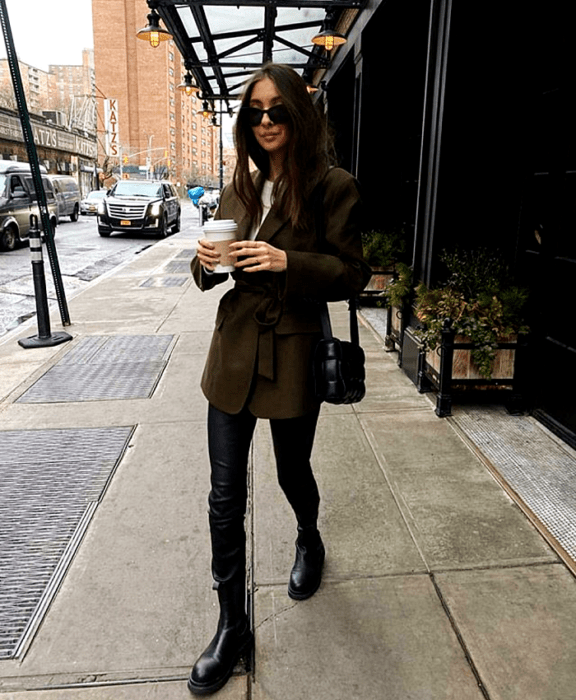 mujer de cabello castaño con lentes de sol, top blanco, abrigo verde militar, leggings negros de cuero, botines negros de piso y bolso mini negro puffer