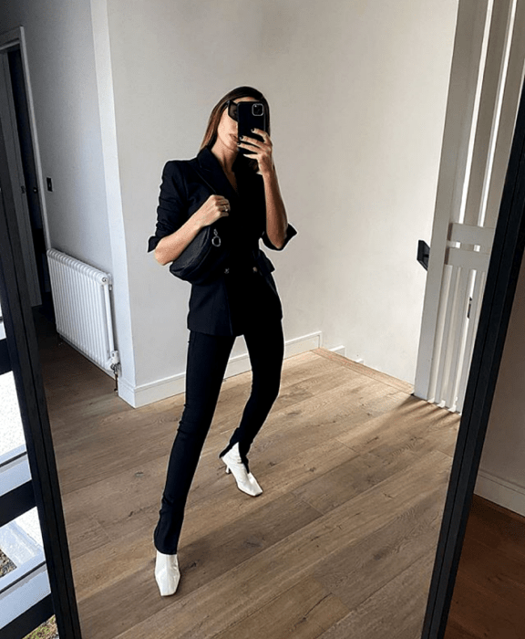 mujer de cabello castaño con lentes de sol, blazer azul marino, leggings negros con abertura en el tobillo, botines blancos de tacón y bolso mini negro