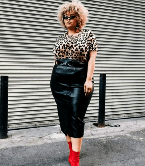 chica morena curvy de cabello rubio usando lentes de sol, camiseta de animal print ajustada de manga corta, falda de cuero midi y botines rojos