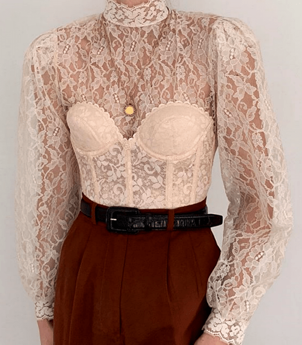 blusa de encaje color crema de cuello alto y manga larga con corsét beige de encaje con cinturón negro de piel y pantalón de vestir naranja oscuro