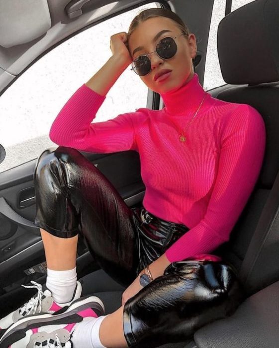 Chica posando dentro de un automóvil con pantalón de látex negro y blusa de cuello alto rosa neón