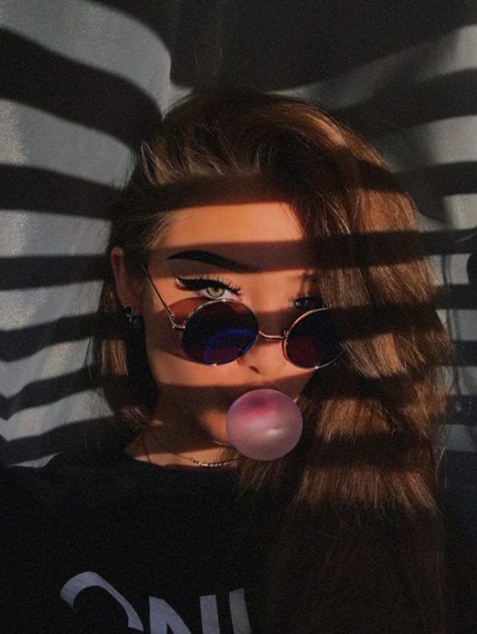 Chica rubia con ojos azules hace burbujas de chicle frente a la ventana y los rayos del sol se reflejan en su cara