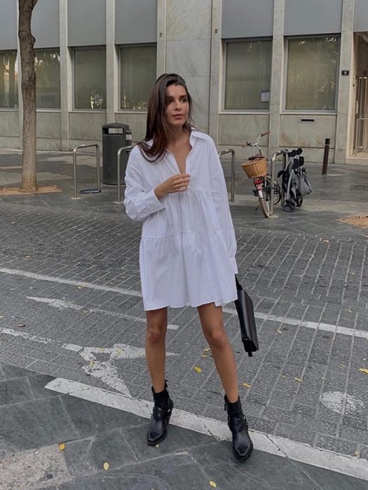 Chica con vestido blanco largo, o camisa de vestir oversized, botas negras y cabello castaño, bolso de mano negro