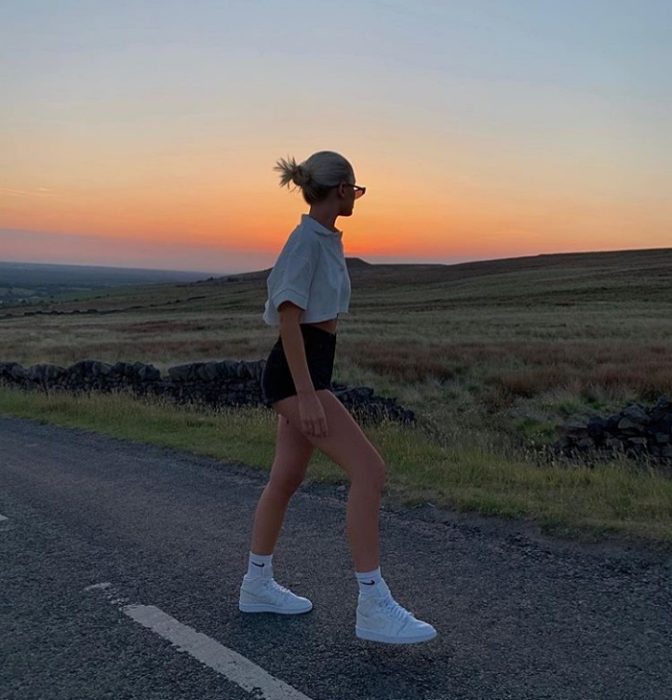 Chica con cabello rubio usando short shorts con una camiseta blanca, tenis deportivos blancos y lentes de sol