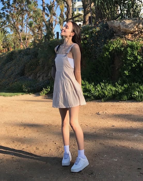 Chica de cabello castaño con un vestido tipo overol color beige, camiseta blanca y tenis blancos