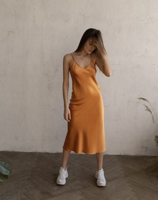 Chica de cabello castaño usando un vestido tipo slip de satén anaranjado con tenis blancos deportivos