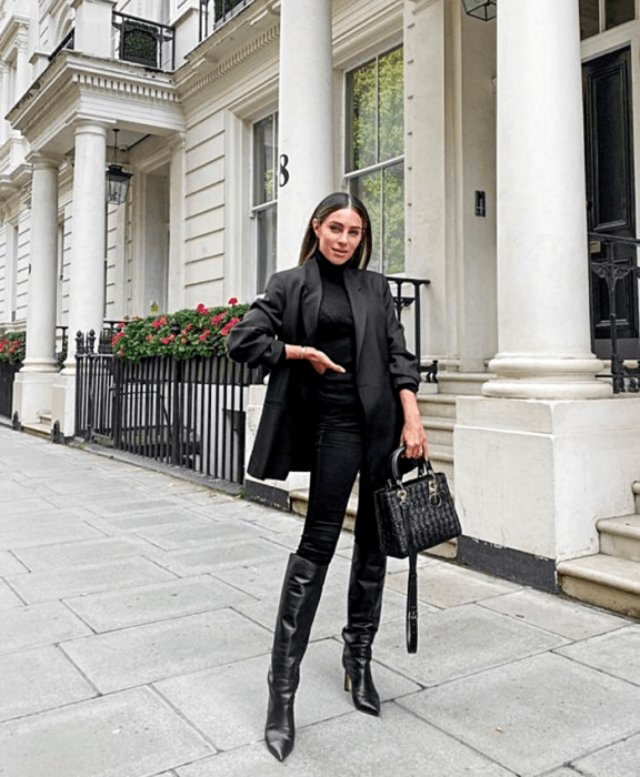 chica de cabello oscuro usando un top negro, saco negro, skinny jeans negros, botas largas de cuero con tacón, y bolso de mano negro