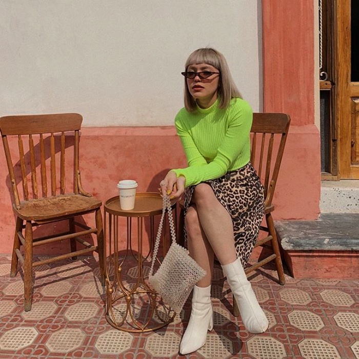 chica de cabello corto con lentes de sol, camiseta de manga larga color verde neón, falda de flores y botas blancas sentada tomando un café