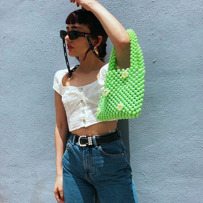 chica con cabello negro, lentes de sol usando un top blanco de manga corta, jeans highwaist y un bolso de cuentas verdes