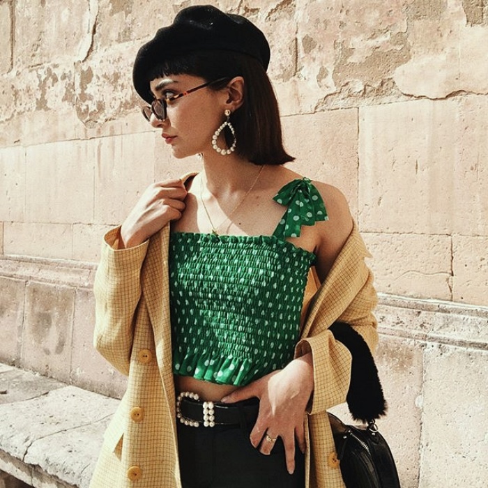 chica de cabello castaño corto usando una boina negra, lentes de sol, top de tirantes color verde esmeralda con puntos blancos, suéter beige, pantalones negros y cinturon con hebilla plateada
