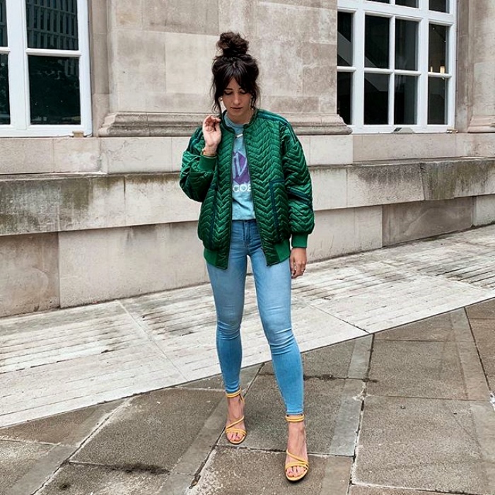 chica con cabello castaño usando un top azul, chamarra bomber jacket color esmeralda y jeans de mezcilla de tiro alto con tacones nude