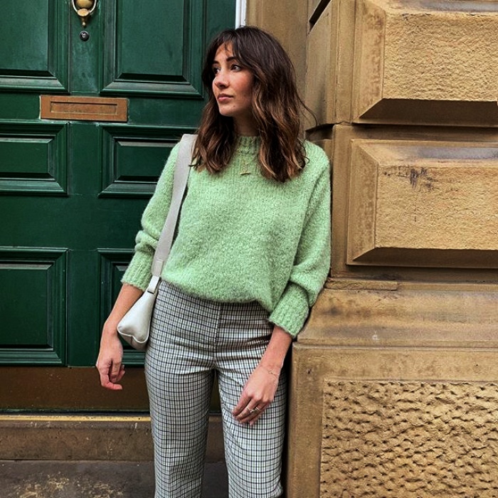 chica de cabello castaño con suéter color verde pastel, pantalones grises y bolso blanco