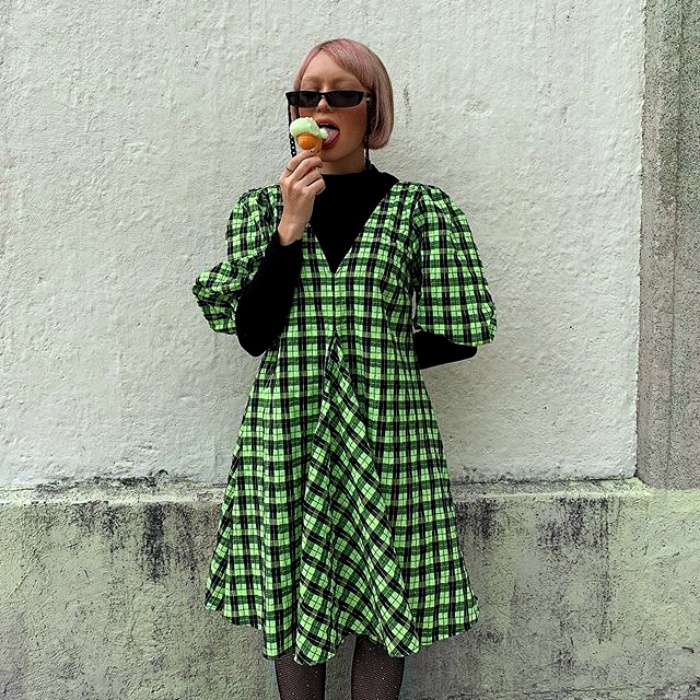 chica de cabello corto y lentes de sol, usando un top de manga larga negro con un vestido verde con cuadros blancos comiendo helado