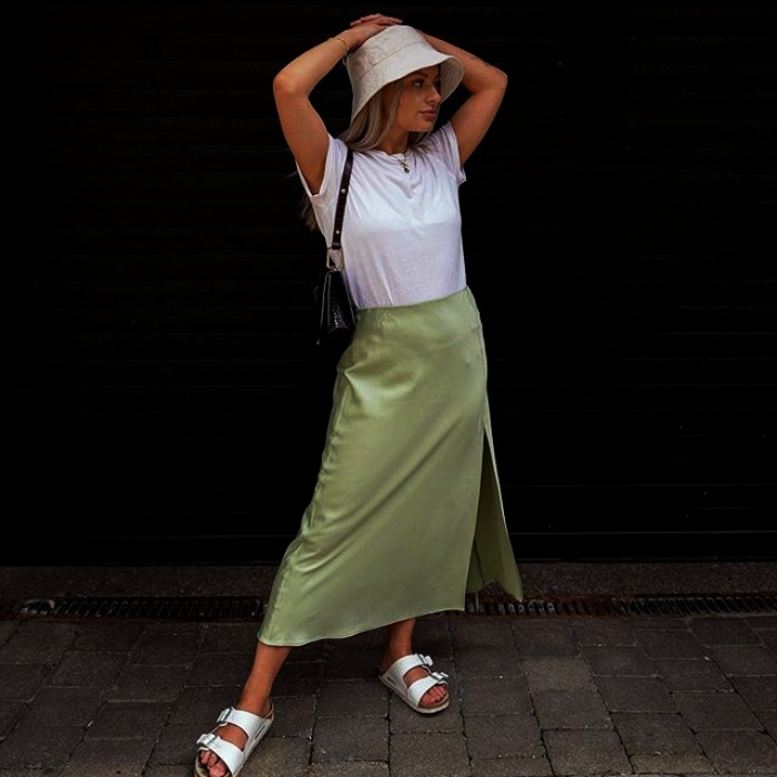 chica rubia con un bucket hat blanco, playera blanca de manga corta, falda de satén verde claro y sandalias blancas