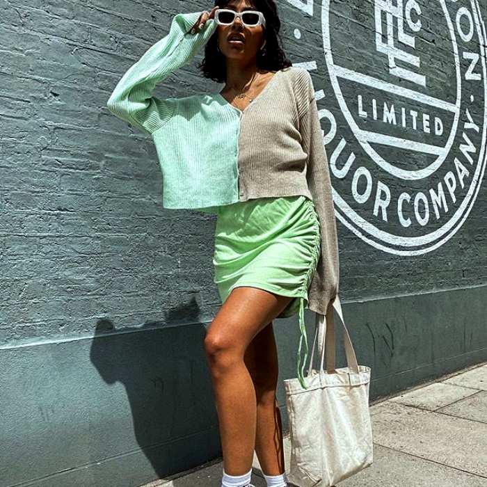 chica morena usando lentes de sol blancos con sueter de dos colores verde menta y gris, usando falda verde de satén y tenis deportivos blancos