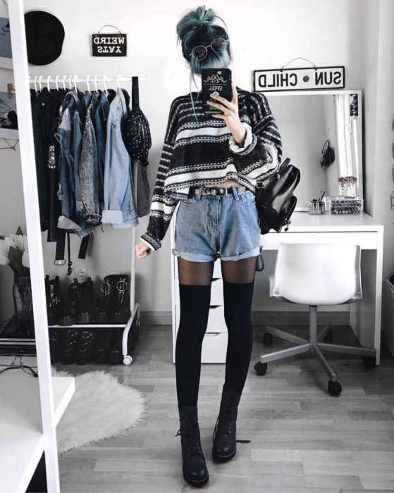 Looks con short vintage; chica tomándose selfie frente al espejo, cabello azul con raíces negras, suéter rayado, medias largas y botas Dr. Martens