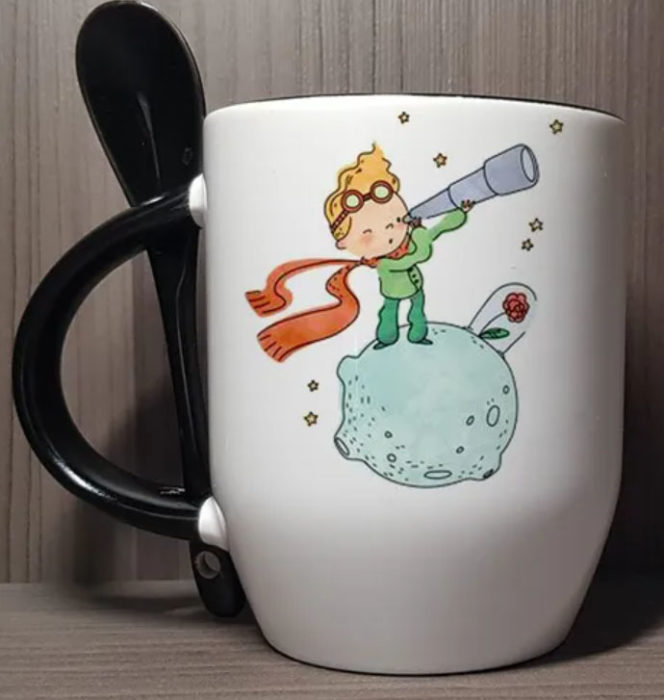 Taza inspirada en El Principito, donde se ve al Principito mirando por un telescopio, con su rosa, y la cucharita de la taza es negra
