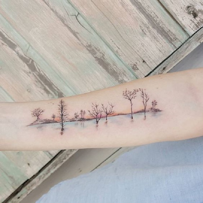 Tatuaje en la zona del antebrazo donde se ve una parte del bosque y el reflejo de un lago en la parte inferior
