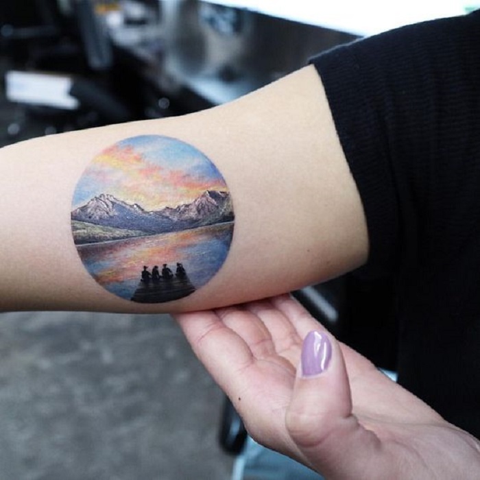 Tatuaje en la parte interna del brazo de un círculo, donde se muestra un muelle con algunas personas sentadas ahí mirando el mar